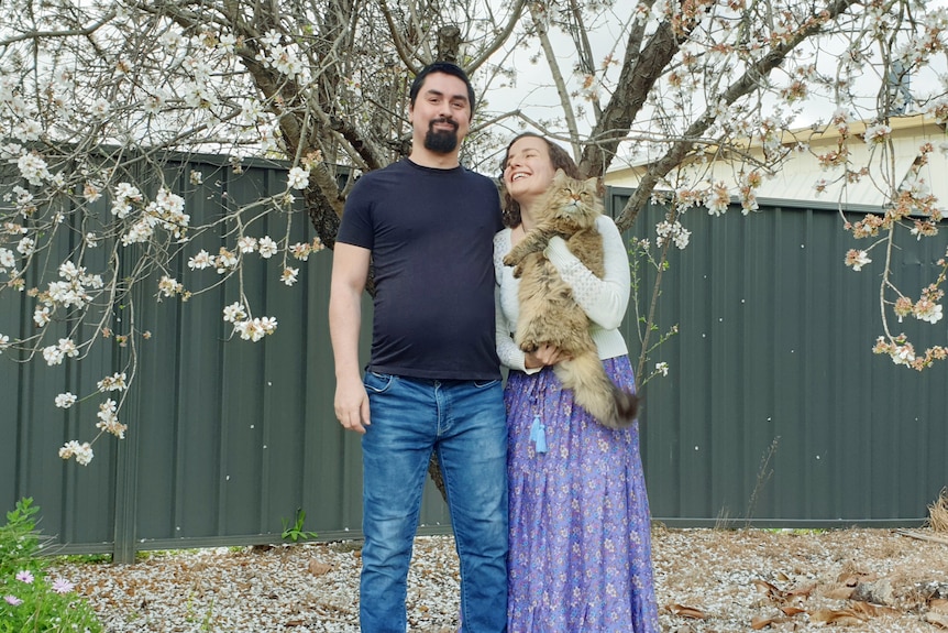 A man and woman standing together, the woman is holding a cat and smiling at the man.