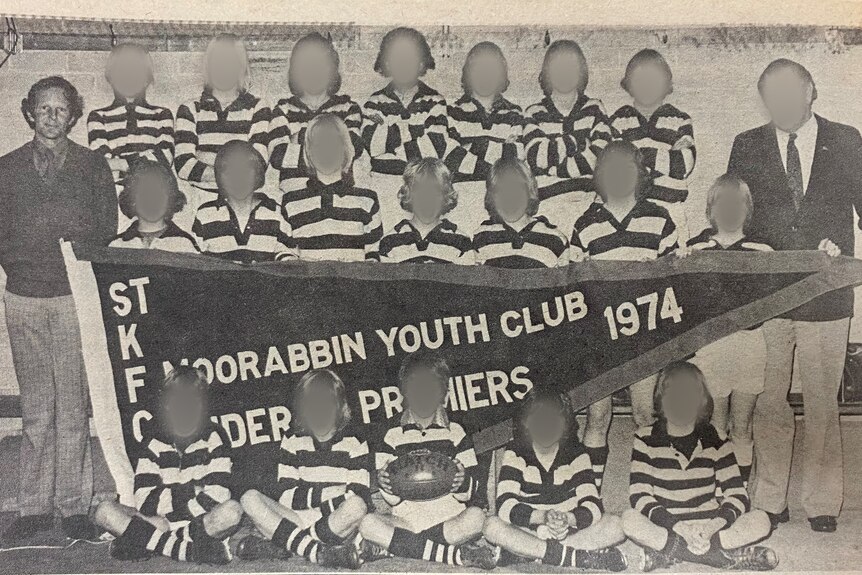 A group of boys pose for a photograph with a pennant, their coach stands to the side.