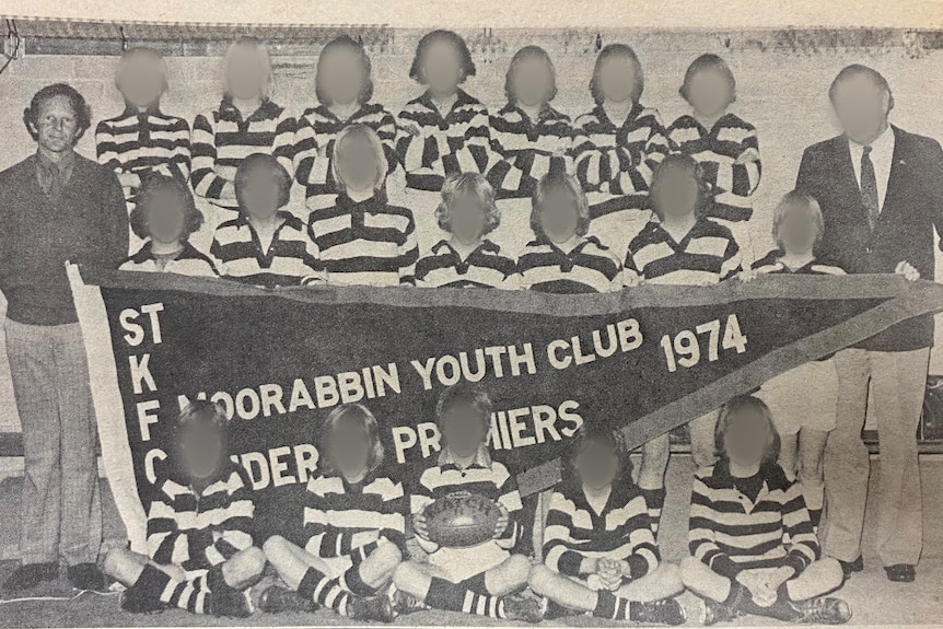 A group of boys pose for a photograph with a pennant, their coach stands to the side.