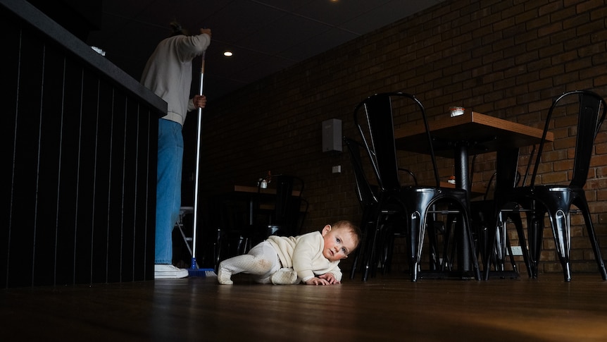 A small baby rolls around on a wood laminate floor as a woman sweeps behind her.