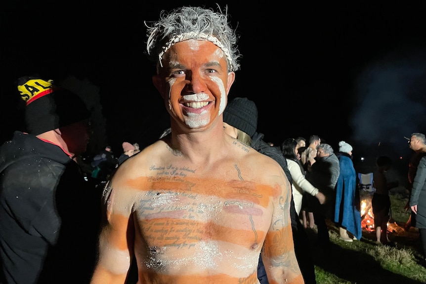 An Aboriginal man with traditional paint and a band around his head smiles at the camera.