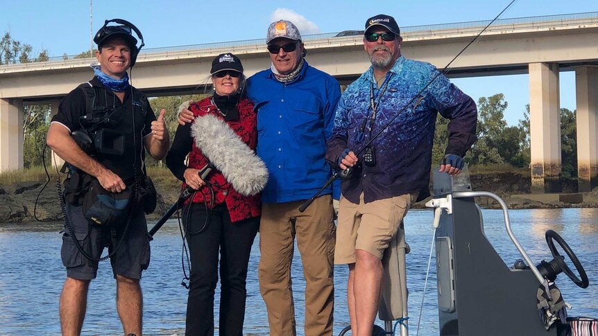 Four people standing on a boat in a river holding fishing rods and TV filming equipment