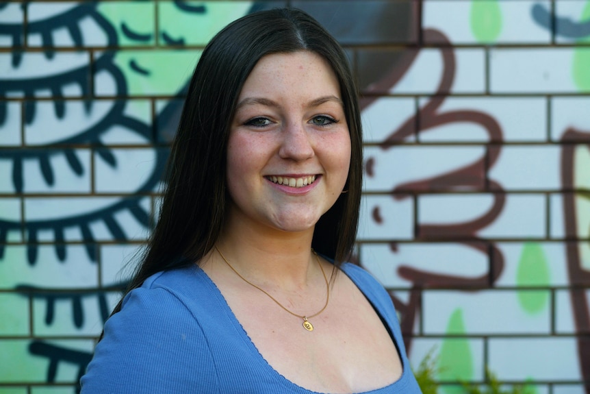 A white skinned woman with brown hair stands in front of a wall of graffiti, wearing a blue top, smiling.