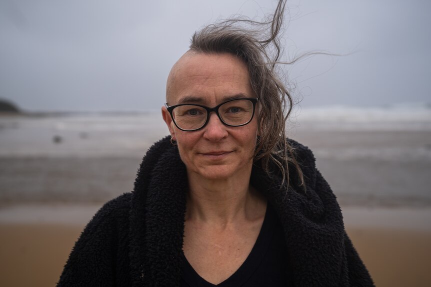 A woman with half a shaved head smiles at the camera on a windy day, the beach is behind her.