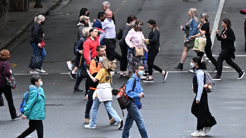 Many people, some wearing face masks, walk across a road.