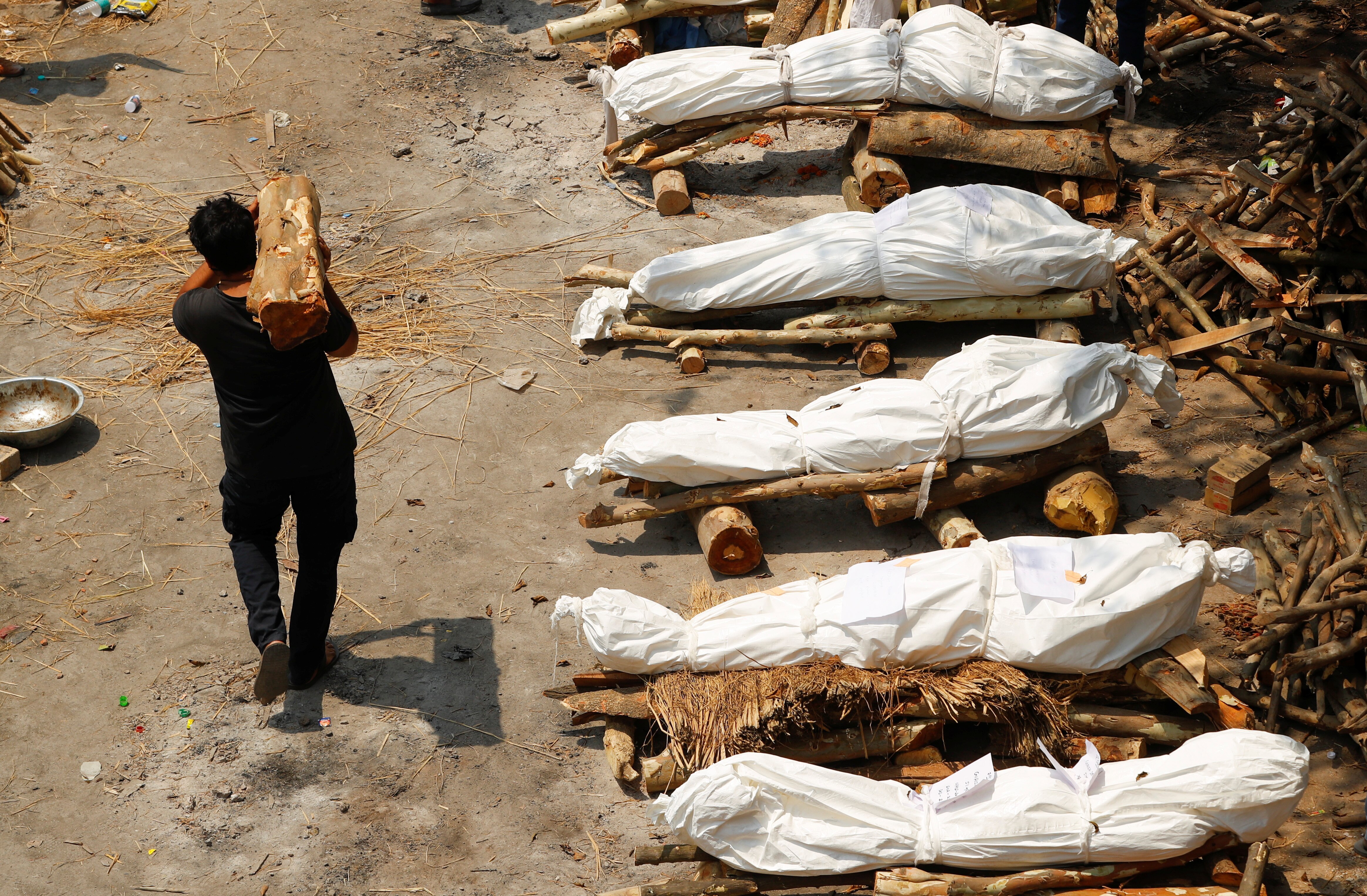 A man carries firewood past bodies of COVID-19 victims wrapped in sheets, waiting to be cremated.