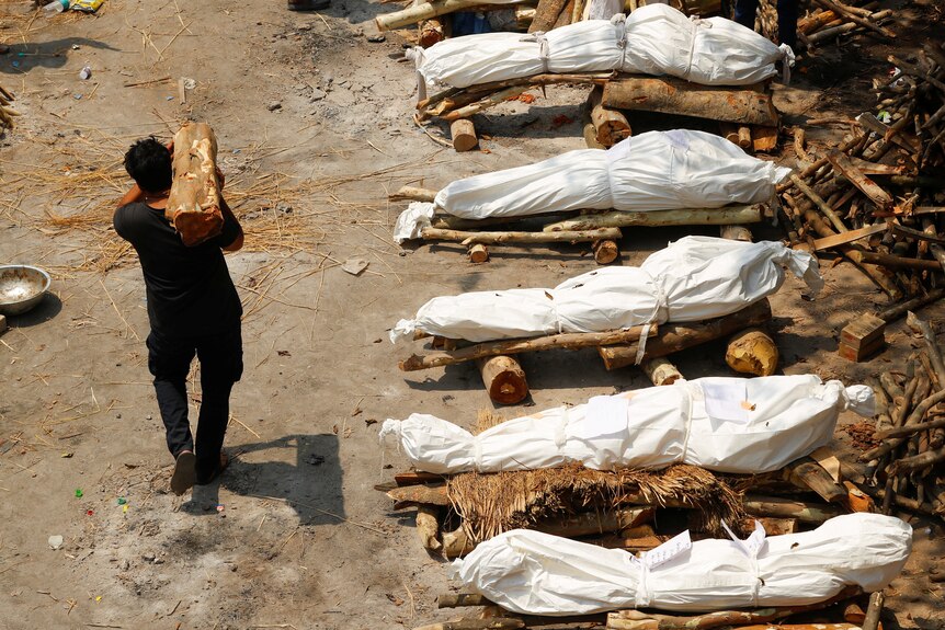 A man carries firewood past bodies of COVID-19 victims wrapped in sheets, waiting to be cremated.