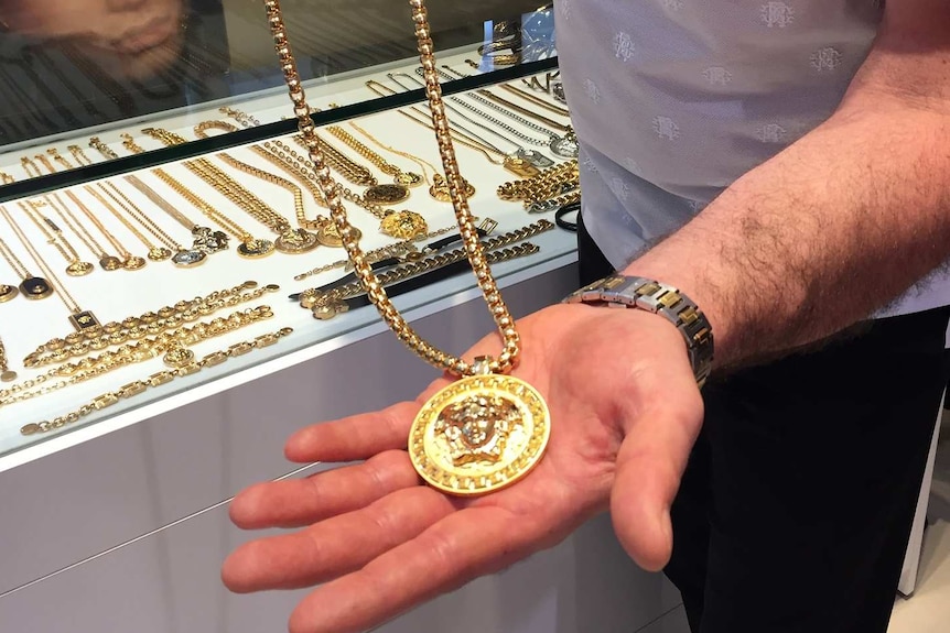 A man holds a gold necklace that has a large golden disc on it in front of a jewellery cabinet.