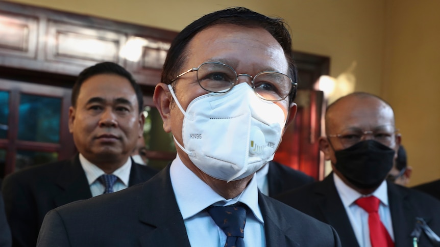A bespectacled and suited man wears a white face mask as he speaks to media outside a courthouse.