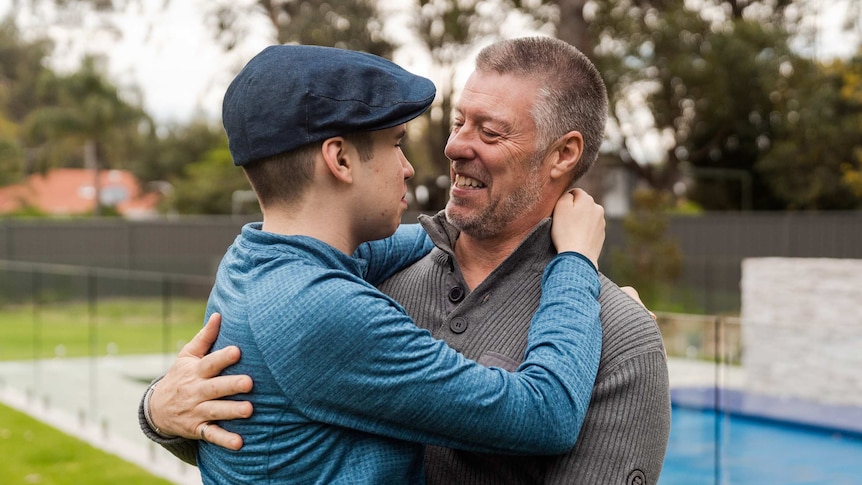 A teenage boy in a blue shirt and flat cap stands hugging his smiling father outdoors.