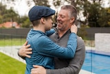 A teenage boy in a blue shirt and flat cap stands hugging his smiling father outdoors.