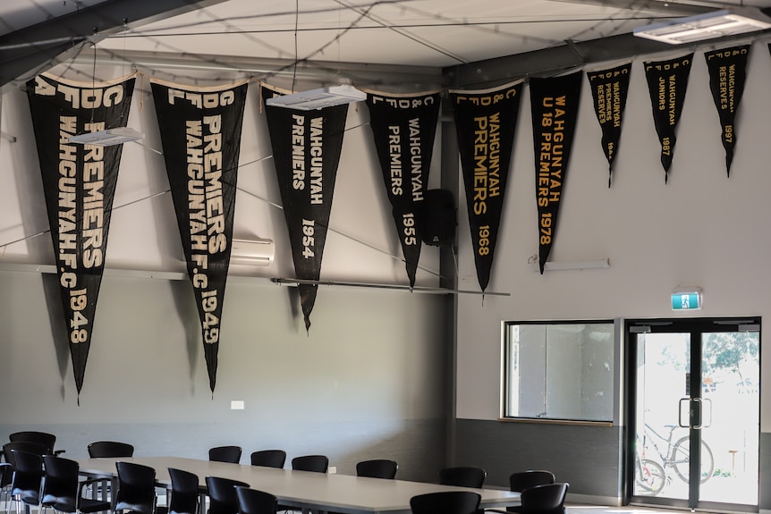 Large pennants displaying text of Premierships from decades ago hang down from the ceiling inside a large white room with tables