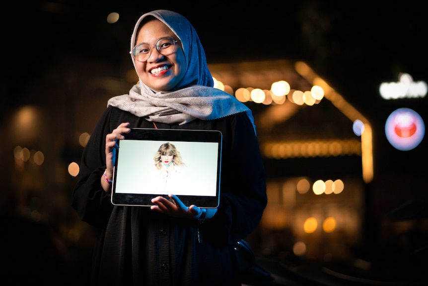 A woman wearing a hijab holds an iPad with a portrait of Taylor Swift infront of a well-lit building at night.