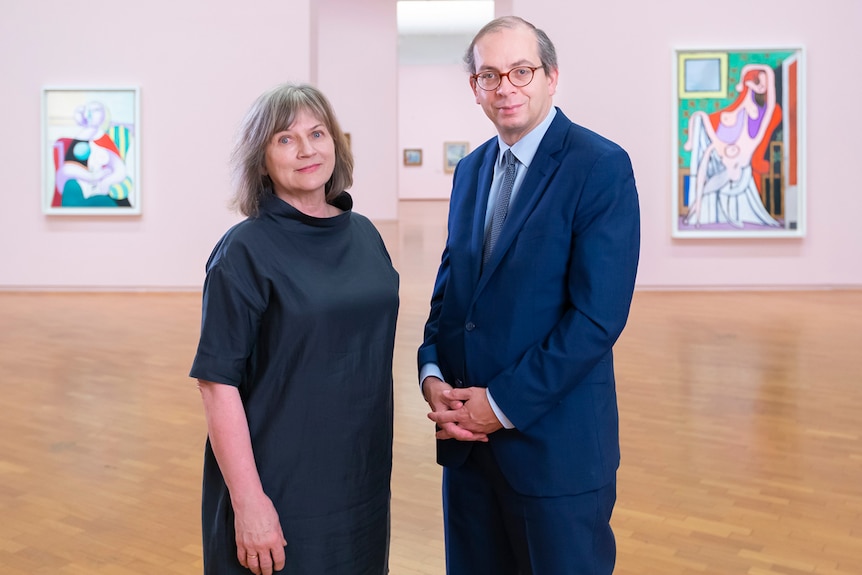 Middle-aged woman with shoulder length grey brown hair next to middle-aged balding man in blue suit, in gallery with pink walls.