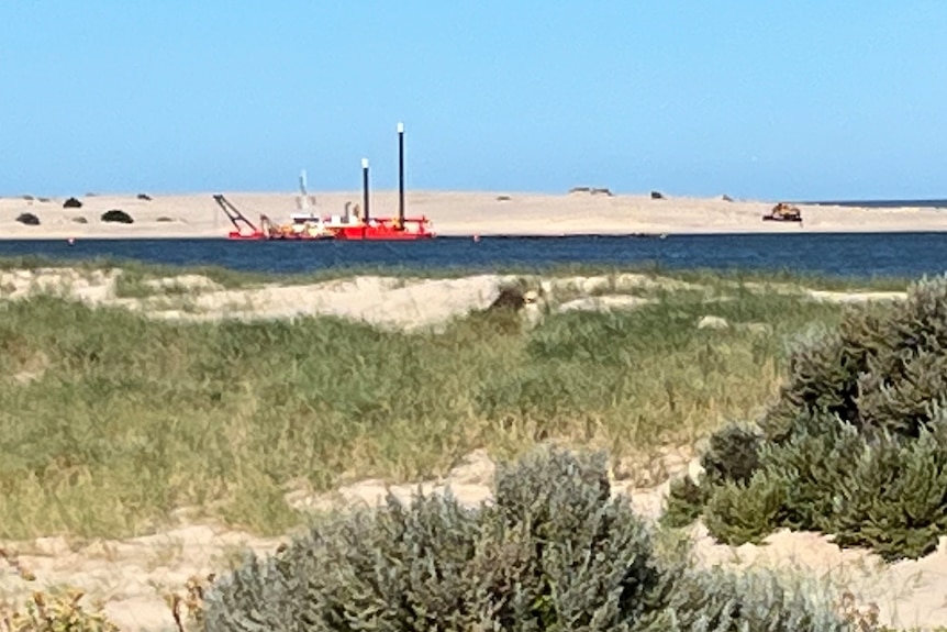 A sign indicating the Coorong National Park stands in front of sand dunes and the mouth of the River Murray and a nearby dredge