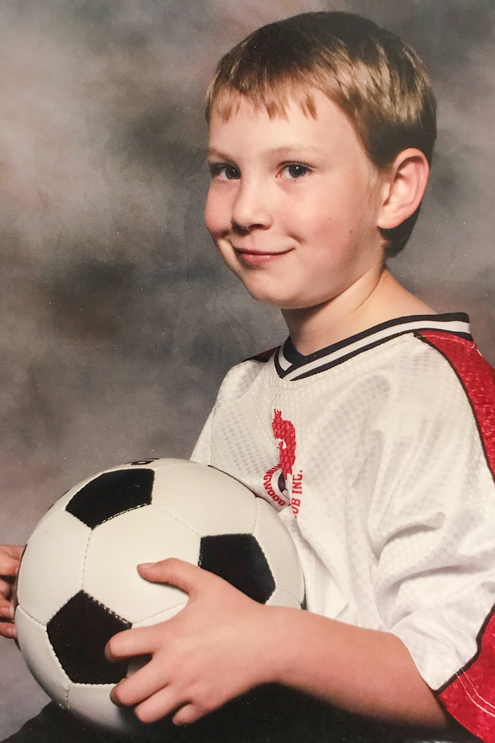 A young Josh sits for a 90s-style portrait, looking off to the side, in a red and white soccer guernsey, holding a soccer ball