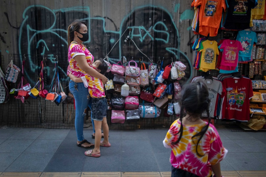 A child hugs her mother as a sibling watches at their sidewalk shop aimed for tourists in Bangkok.