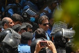 Imran Khan wears sunglasses as he is ushered through a crowd surrounded by armour-wearing police officers on city street