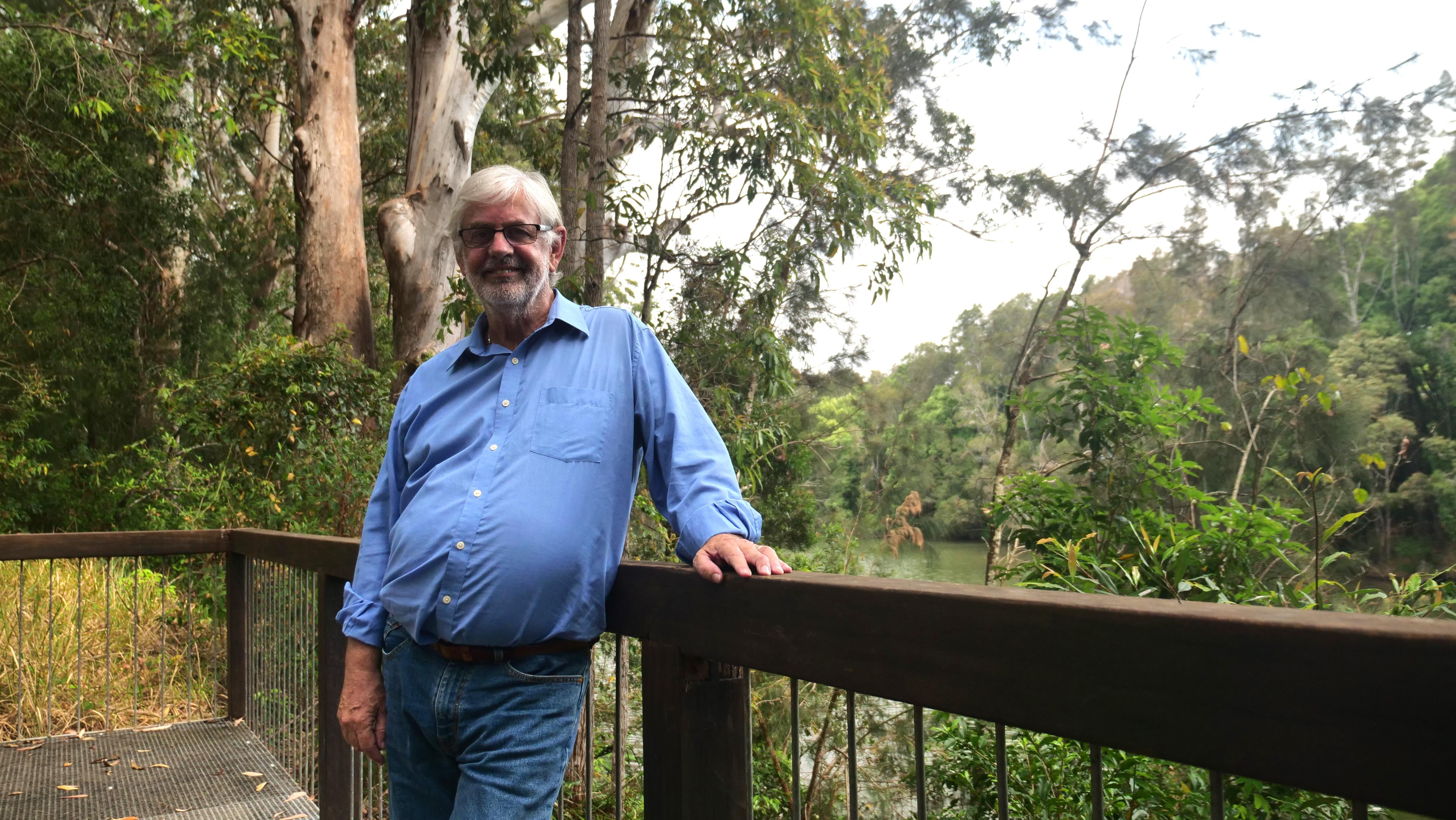 Um homem de camisa azul de mangas compridas e jeans está encostado em uma grade sorrindo com árvores nativas e um riacho ao fundo.