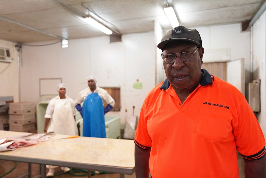 PNG man wearing a hi-vis orange shirt, glasses and a black cap with two assistants behind him.