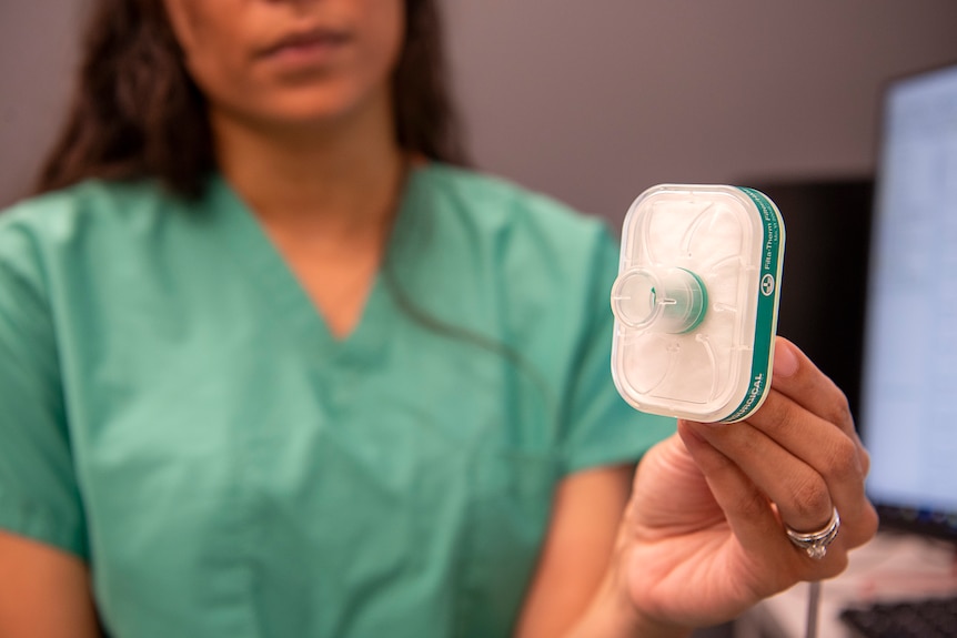 A woman in green scrubs holds up a device to the camera. It has a mouth piece connected to a rectangular filter with a tube