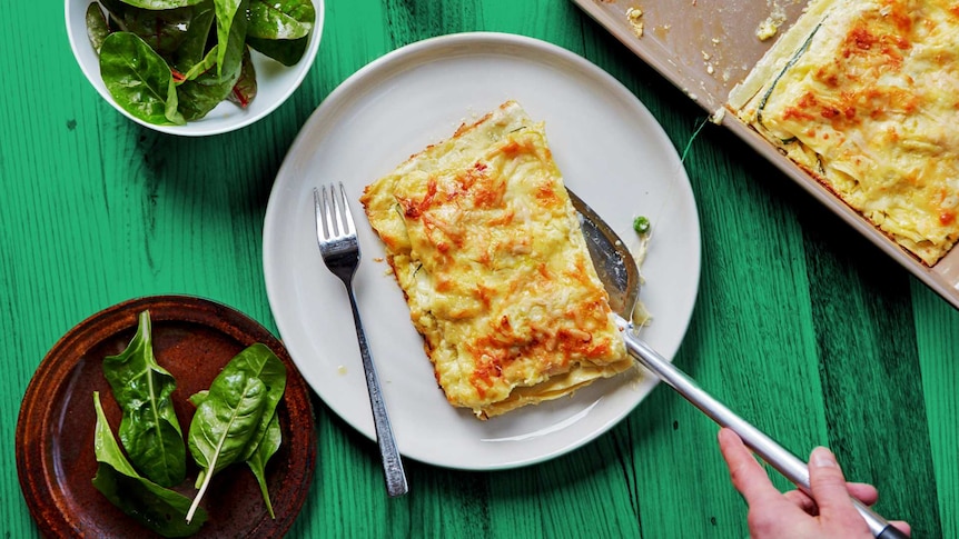 A slice of vegetable lasagne is placed on a pate with a metal spatula, a meat free dinner with a green side salad.