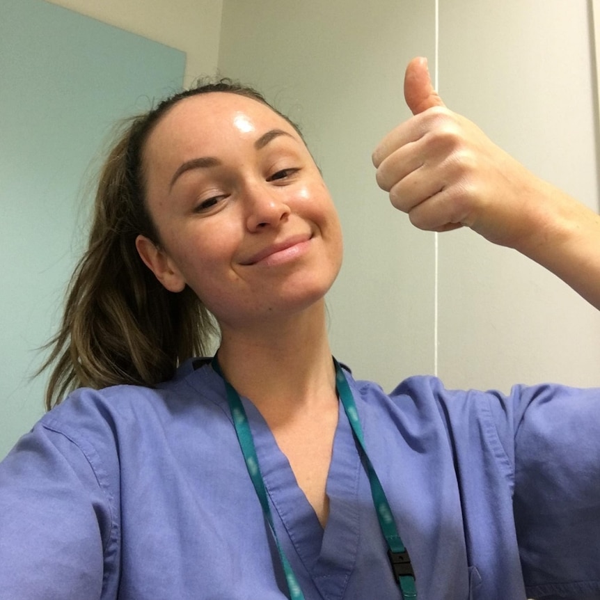 A nurse wearing blue scrubs leans back from the camera to give a thumbs up and tired smile