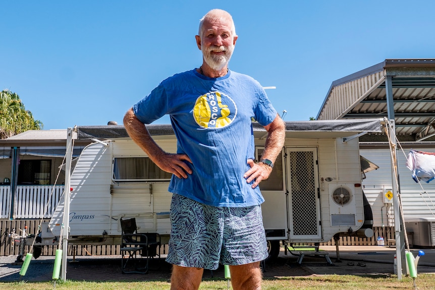 A man stands in front of a caravan in a yard smiling.