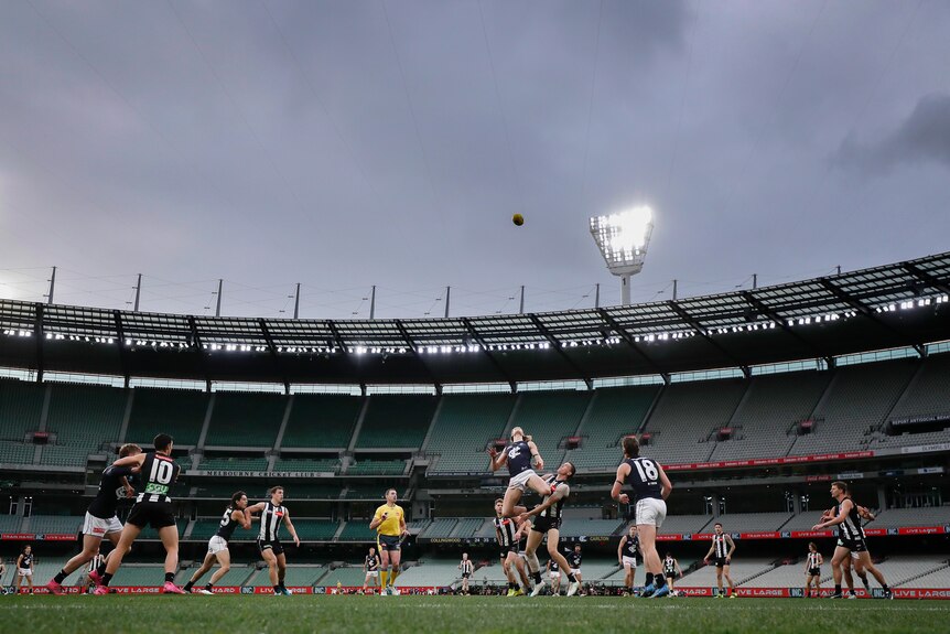 La pelota vuela alto cuando los jugadores de dos equipos de la AFL saltan a la competencia, con un reflector y gradas vacías en el fondo.