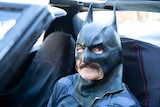 A man in a rubber Batman mask sits in a dark car.