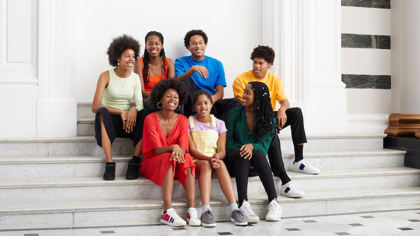 Seven musicians from the Kanneh-Mason family sit together smiling on some marble stairs.