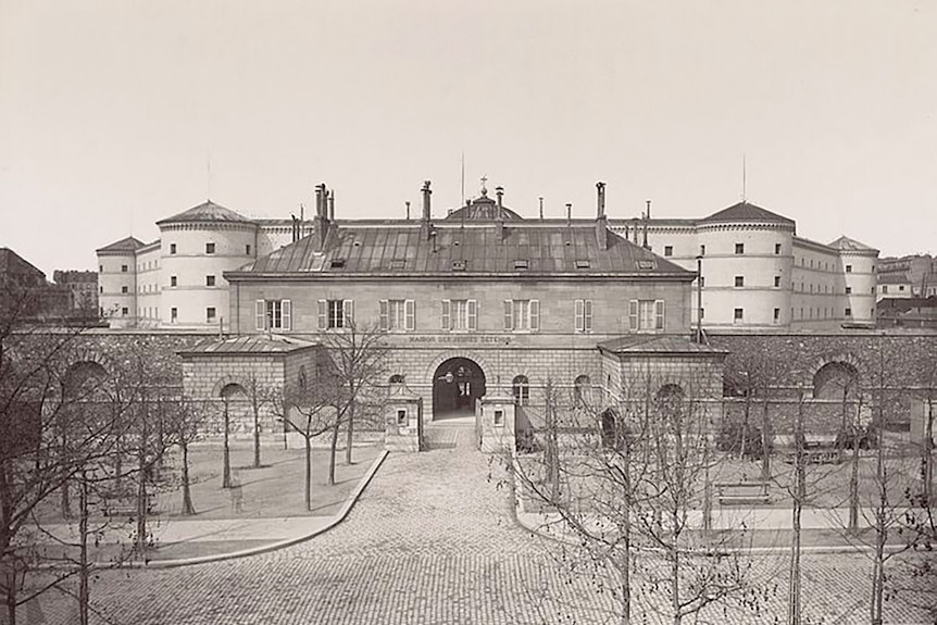 Black and white photo of a building with tall stone fences, an arched front entrance and large courtyard.