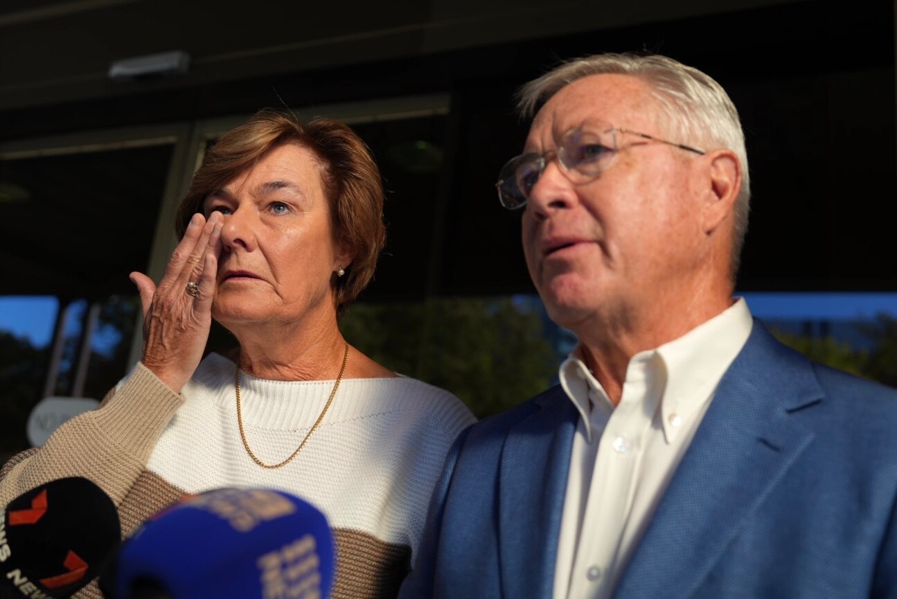 A woman wipes away a tear next to a man in front of media microphones