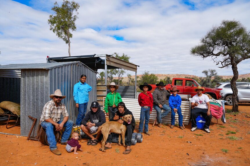 Group shot of stockmen and boys in a dusty red dirt paddock with a tin shed.