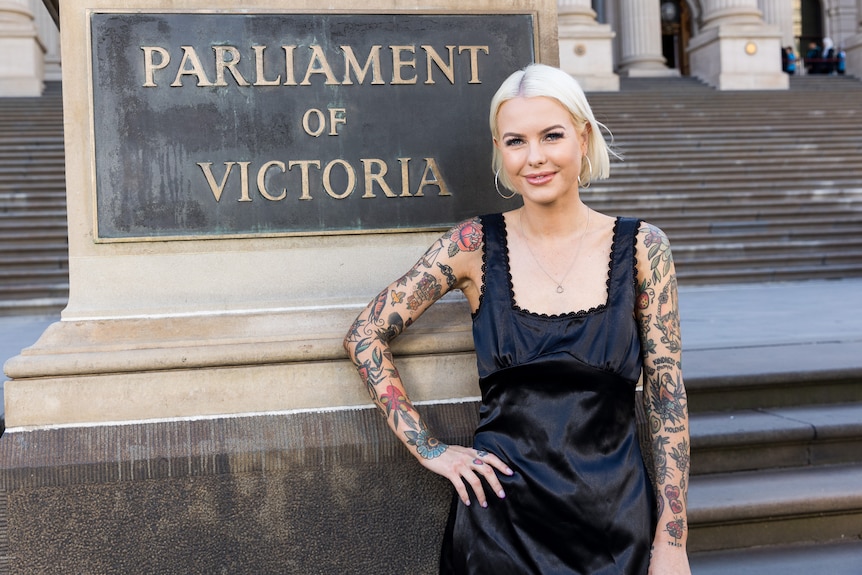 Women with short blonde hair and tattoos on her arms stands in front of a Parliament of Victoria sign and smiles.