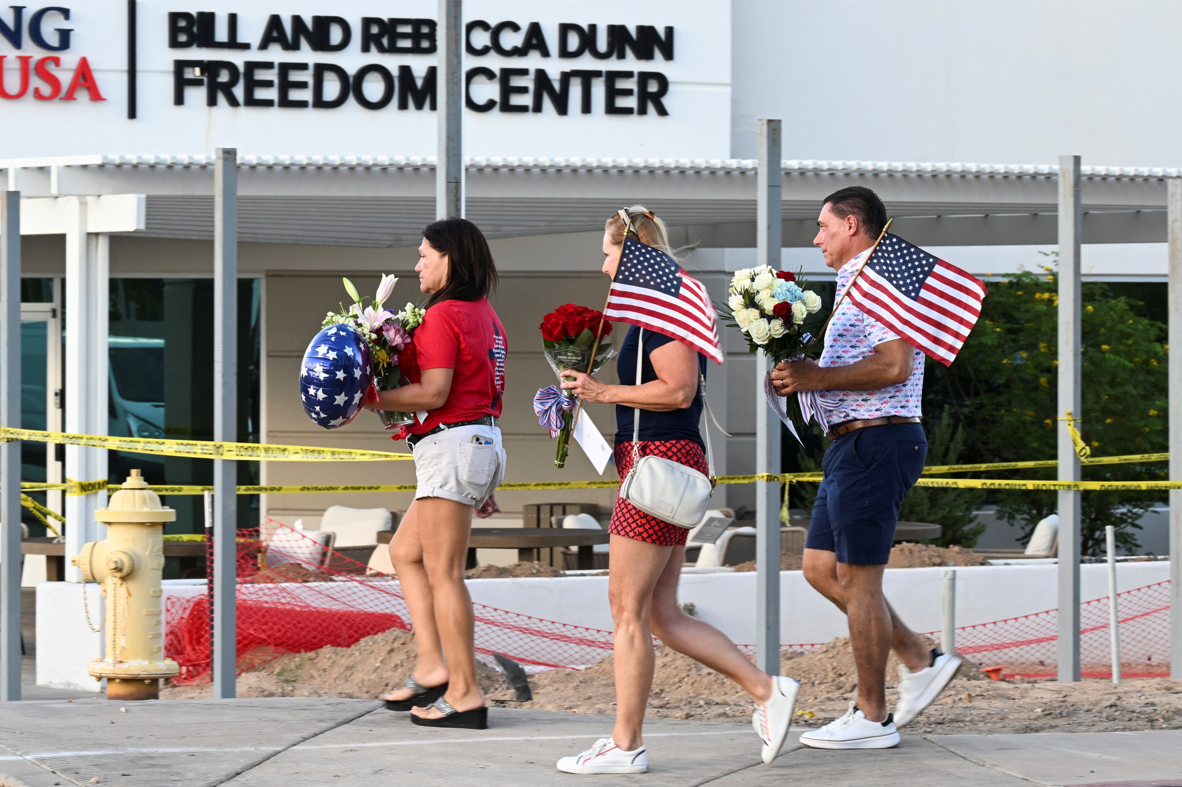 Three people carry US flags and flowers outside a building that is surrounded by policy tape.