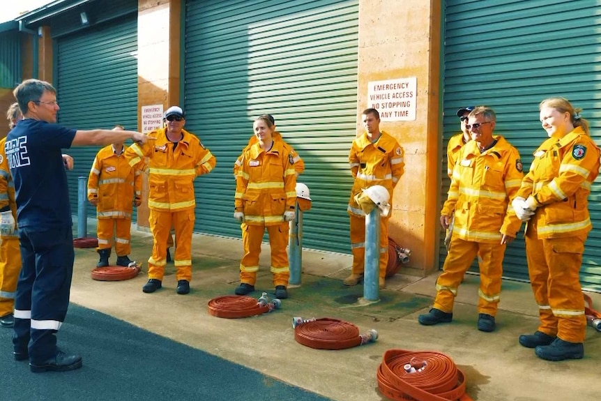 A group of nine people in orange fire gear stand facing a man in blue, who is pointing towards three garage doors.