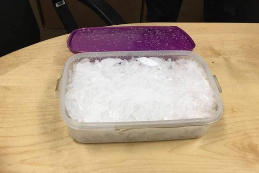 A plastic container filled with ice crystals sits on a wooden table with its purple lid laying behind it.