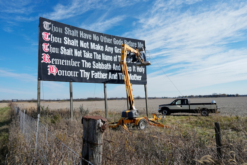 A road sign displaying five of the Ten Commandments sits behind a fence, with a mobile crane on its left.