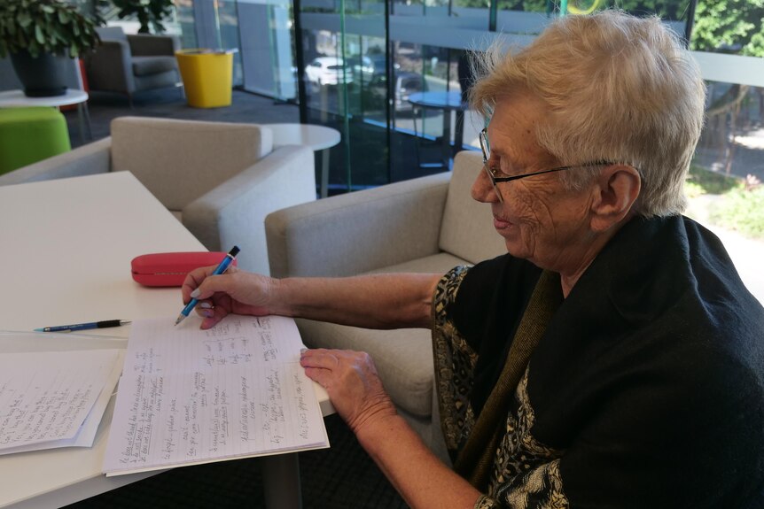 Older woman sitting at a library desk holding a pen and smiling at her page.