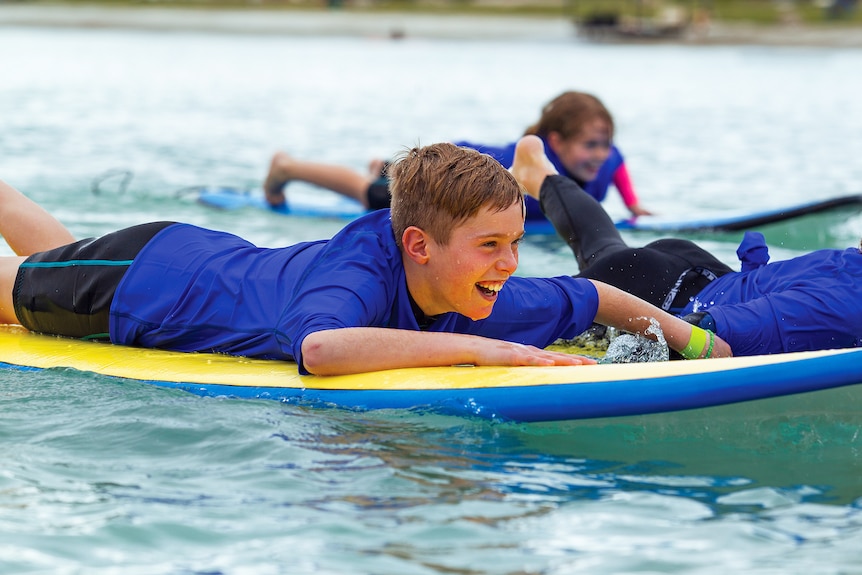 A young boy in a rashie and board shorts lying on a surf board and a young girl behind him paddling on her board