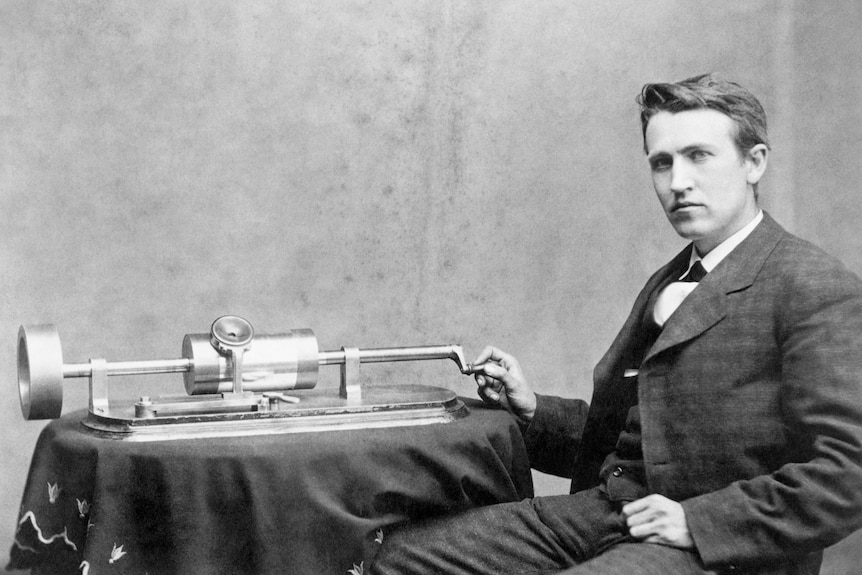 Black and white photo of a young Thomas Edison sitting at a table upon which one of his phonographs sit.