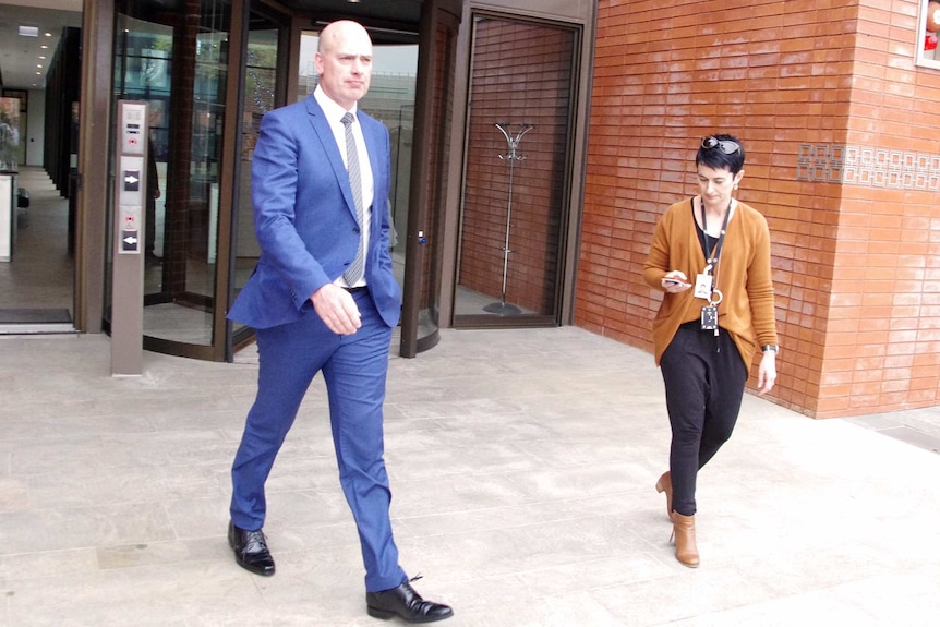 Dean Nalder walks out a door and along a footpath wearing a blue suit, with a woman holding a phone beside him.