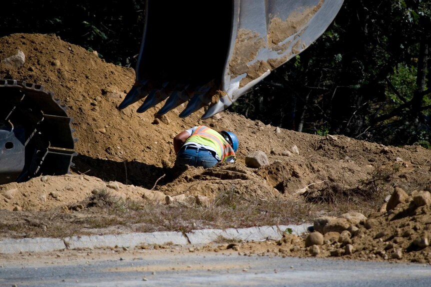 A worker in a blue hard hat and high-vis vest digs in a ditch, excavator scoop in foreground.