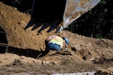 A worker in a blue hard hat and high-vis vest digs in a ditch, excavator scoop in foreground.