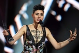 A woman speaks into a microphone while holding an aria award. On her chest is written people over profit.
