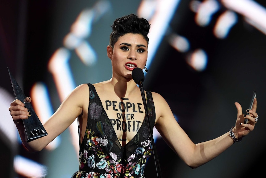 A woman speaks into a microphone while holding an aria award. On her chest is written people over profit.