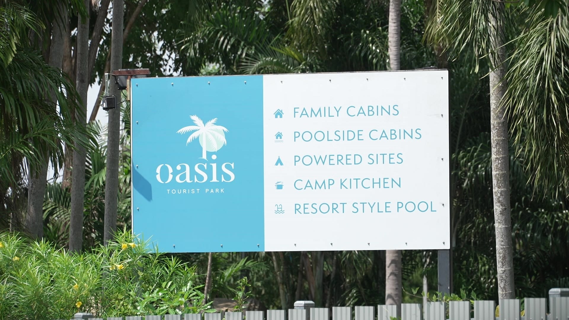 A blue and white sign in among tropical palm trees that reads 'Oasis Tourist Park'.