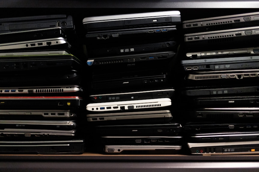 Dozens of laptops stacked on top of each other on a shelf.