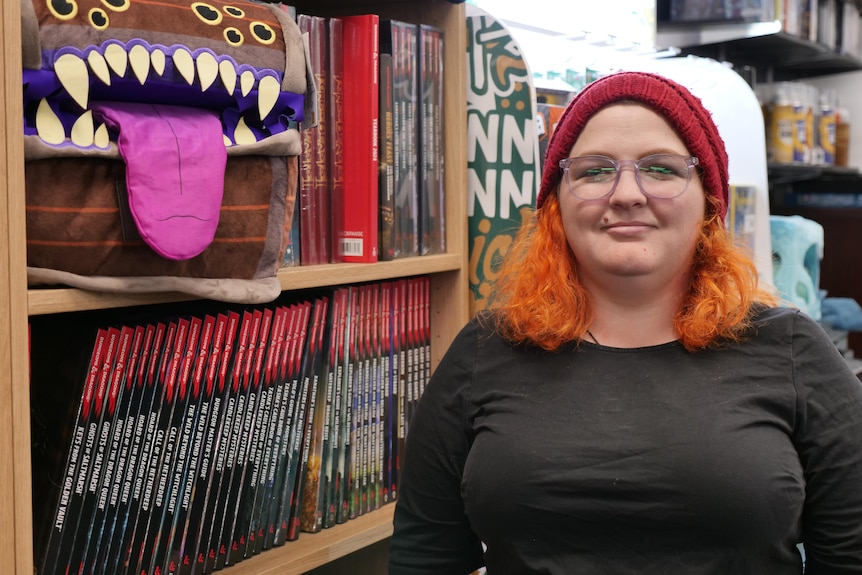 身穿黑色衣服、留着橙色头发、戴着红色帽子的女人站在书架旁，书架上放着一个柔软的宝箱玩具